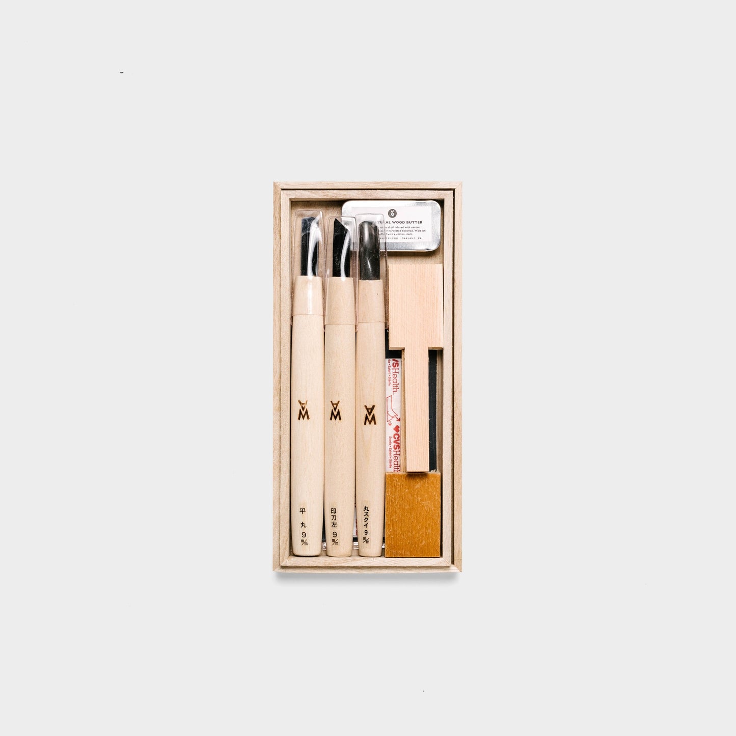 Beginner spoon carving kit shown open, displaying three carving tools, two spoon blanks, sandpaper, wood butter, and a bandaid. By Melanie Abrantes Designs.