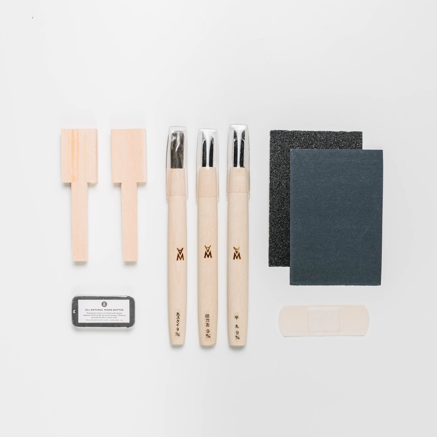 Beginner spoon carving kit items laid out. Two spoon blanks, wood butter, three carving tools, two sandpapers with different grits, and a bandaid. By Melanie Abrantes Designs.