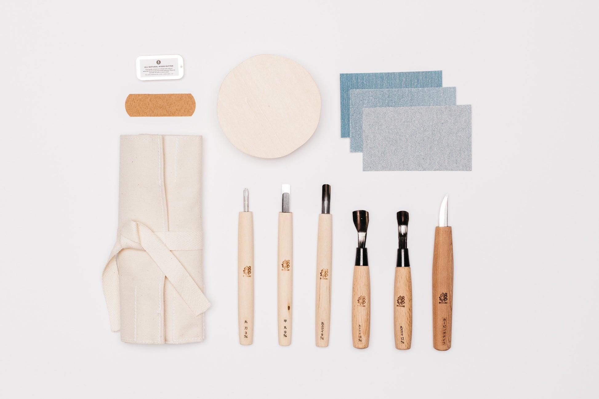 Japanese bowl carving kit. The items in the kit are laid out as follows, MA wood butter, bandaid, beige tool roll, six carving tools, bass bowl blank, and three sandpaper grits. 