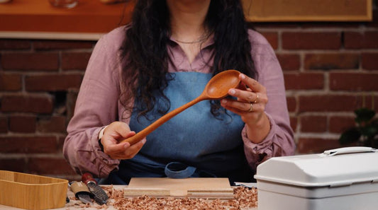 DIY tutorial for carving a cooking spoon. Finished product in hand. Knife, shinto rasp, and bench hook placed on table. Learn safety awareness. By Melanie Abrantes Designs.
