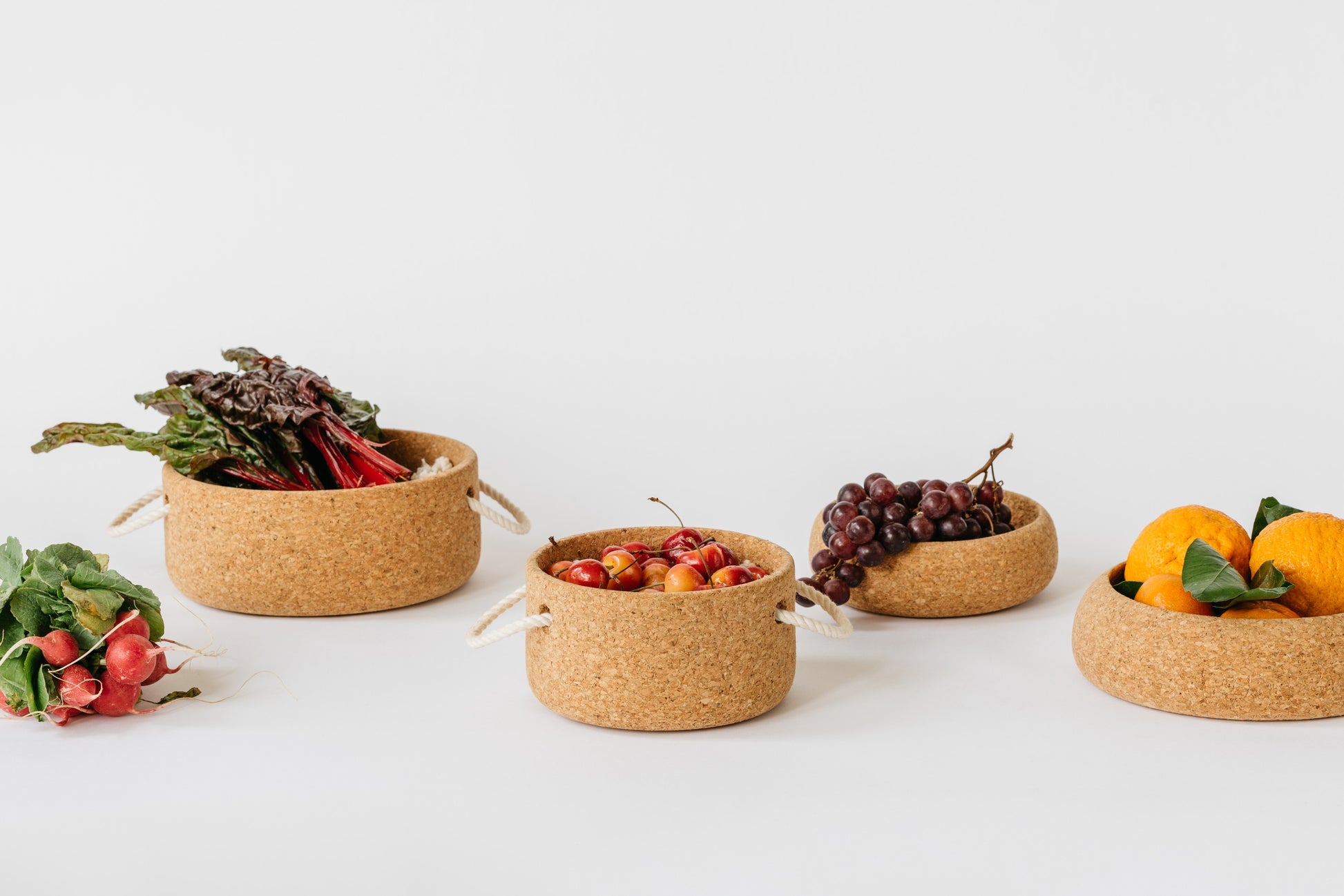 Cork bowls by Melanie Abrantes Designs. Large and small bowls with rope handles. Other cork bowls with no handles. Bowls are filled with fruits and vegetables.