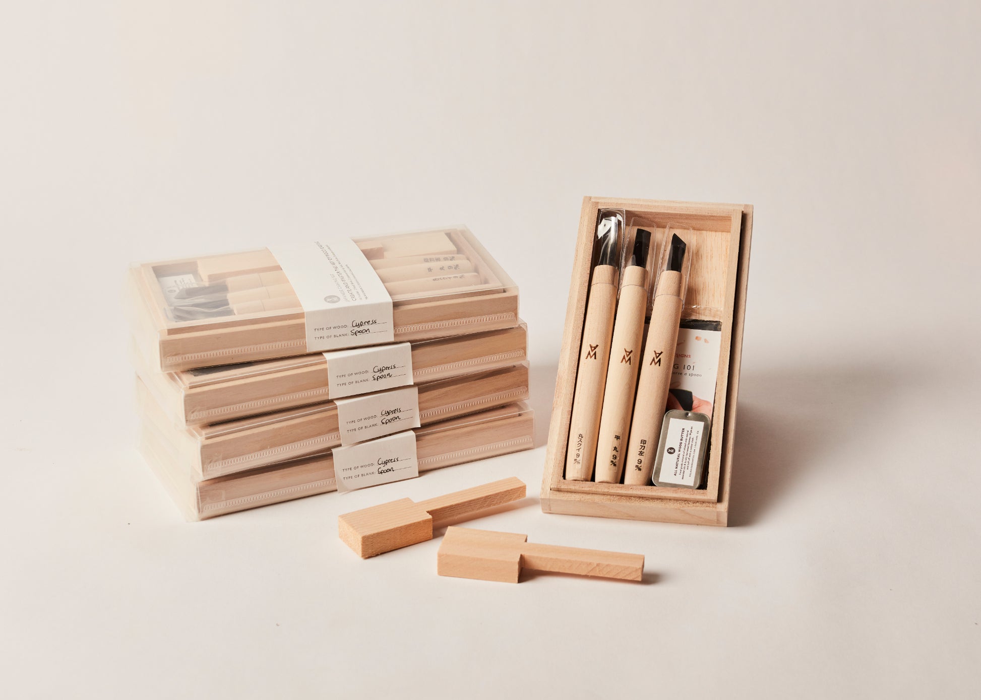 Beginner Spoon Carving Kit. Four kits stacked up with one open kit displaying three carving tools, instructions, sandpaper, bandaid, wood butter, and two spoon blanks. By Melanie Abrantes Designs.