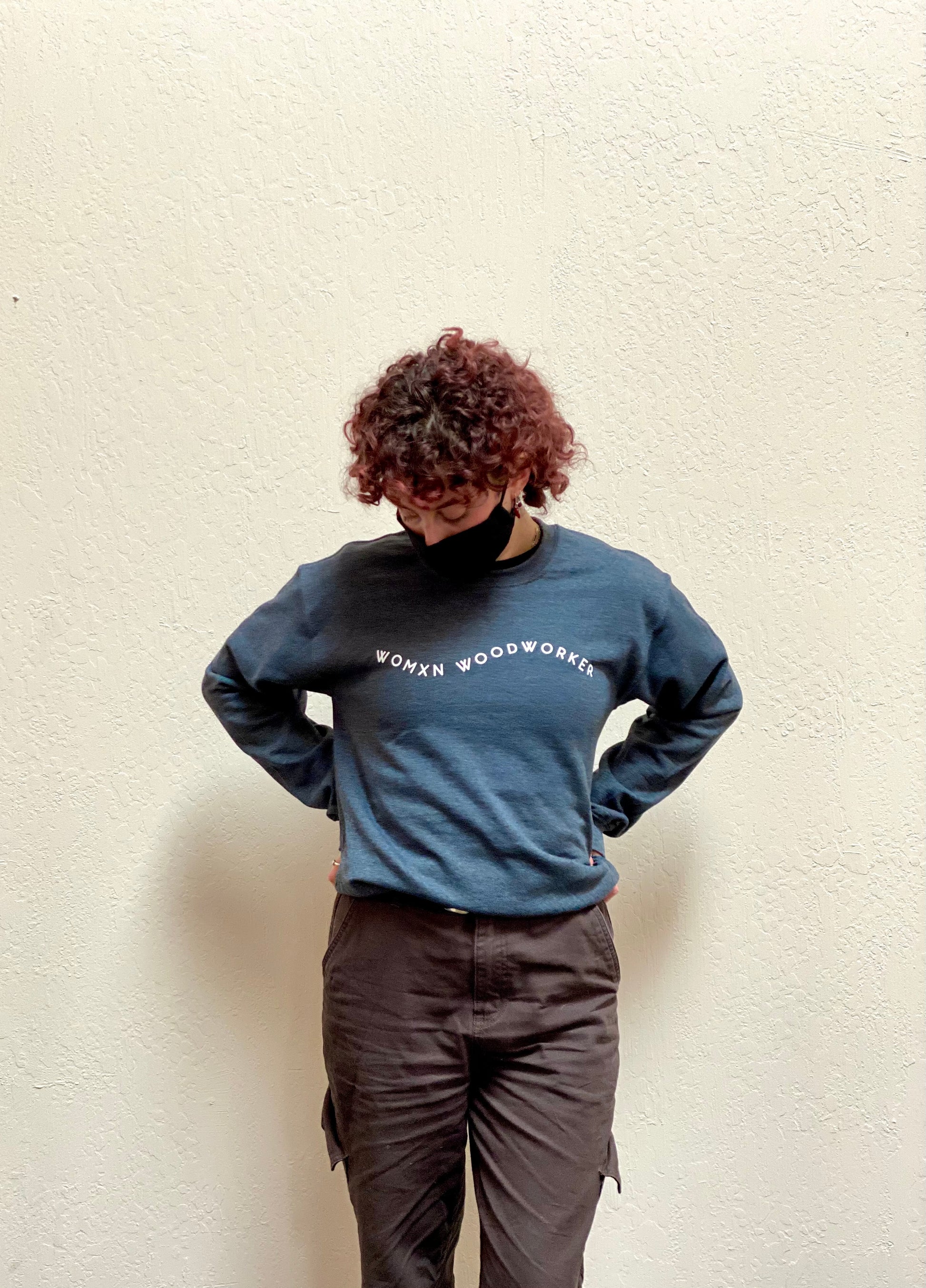 Person with red curly hair wearing Heather grey crew neck sweatshirt. Reads "Womxn Woodworker" in white font.