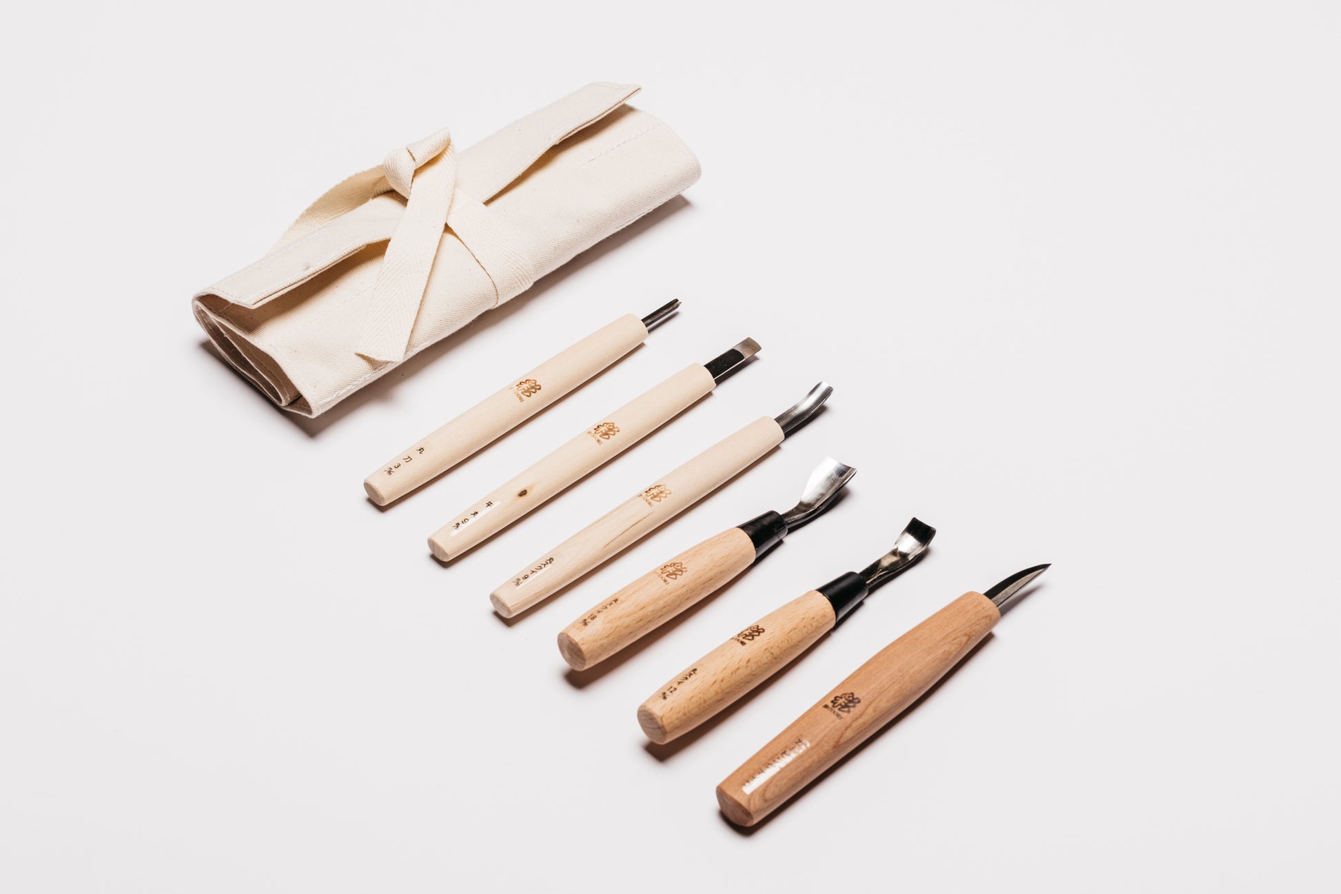 Laid out on a white background is the tool roll tied up and the six carving tools you will use to make your wood bowl. Curated by Melanie Abrantes Designs.